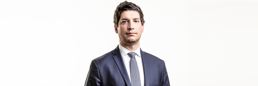 Un uomo in abito scuro e cravatta è in piedi su uno sfondo bianco. Appare sicuro di sé e professionale, trasmettendo un tono formale e serio.